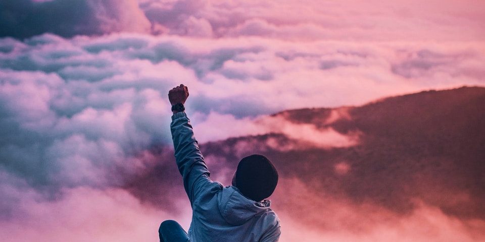 man sitting on mountain cliff facing white clouds rising one hand at golden hour